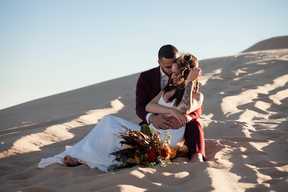 bride and groom sitting in sand dunes in Arizona with wedding bouquet beside her