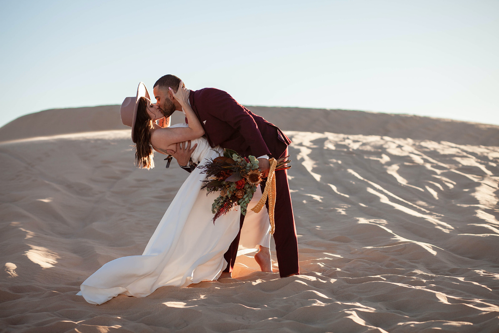 Groom dips bride during kiss in desert elopement with sunset in the background