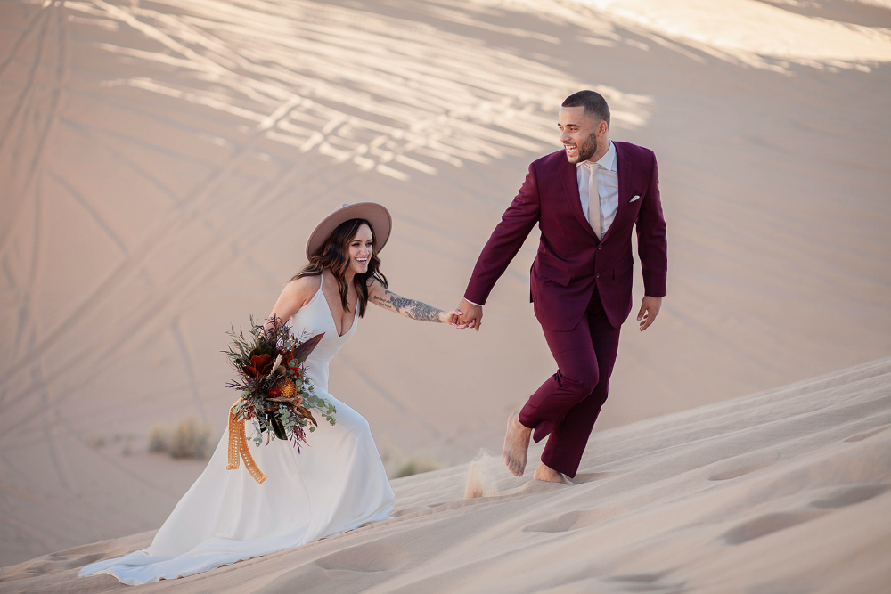 Desert Elopement in Arizona with bright florals and flowy dress climbing up sand hill